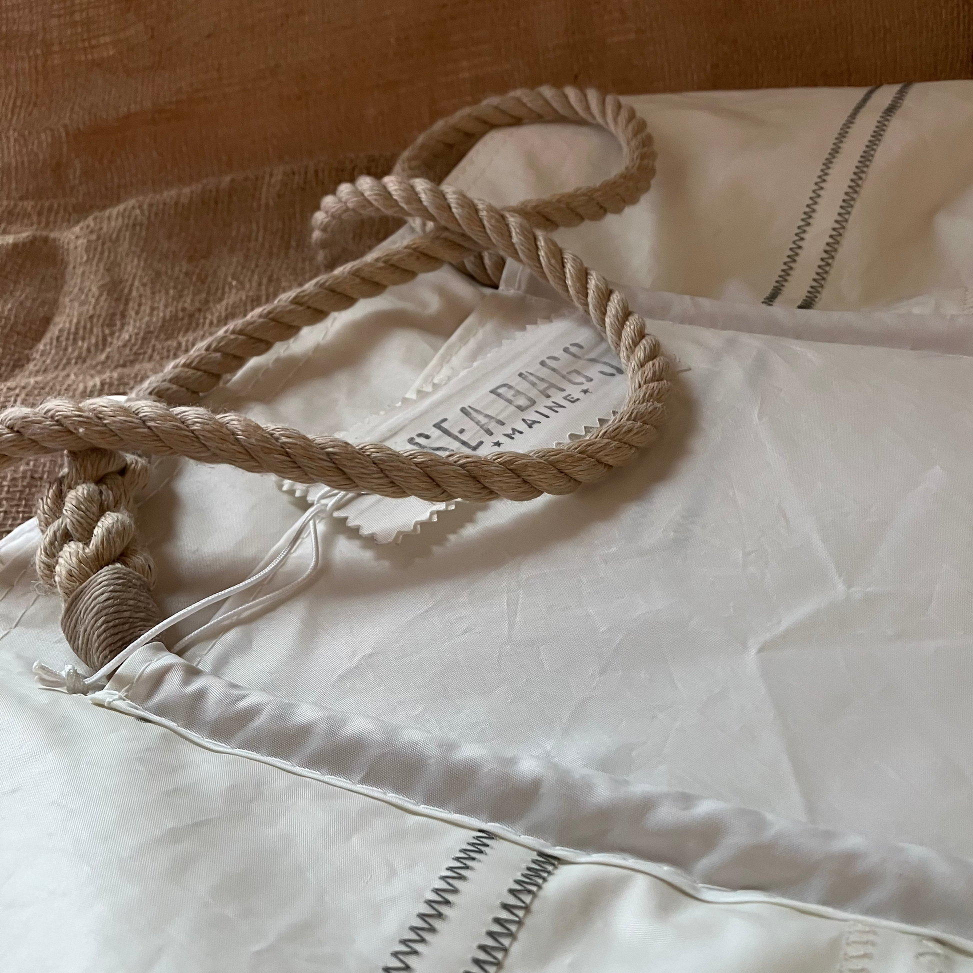 A close-up of the Sea Bags Wharf Tote, made from white sail cloth with rope handles and visible stitching, shown on a beige textured surface.