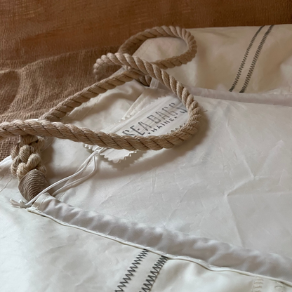A close-up of the Sea Bags Wharf Tote, made from white sail cloth with rope handles and visible stitching, shown on a beige textured surface.