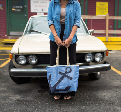 A person holds a Navy Anchor on Chambray Medium Tote, featuring a light blue fabric with a dark blue anchor graphic and thick rope handles, while standing in front of a tan car.