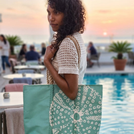 A woman stands by a pool at sunset carrying a Seafoam Sea Urchin Medium Tote, a light green recycled sail cloth bag with a white sea urchin print and rope handles.