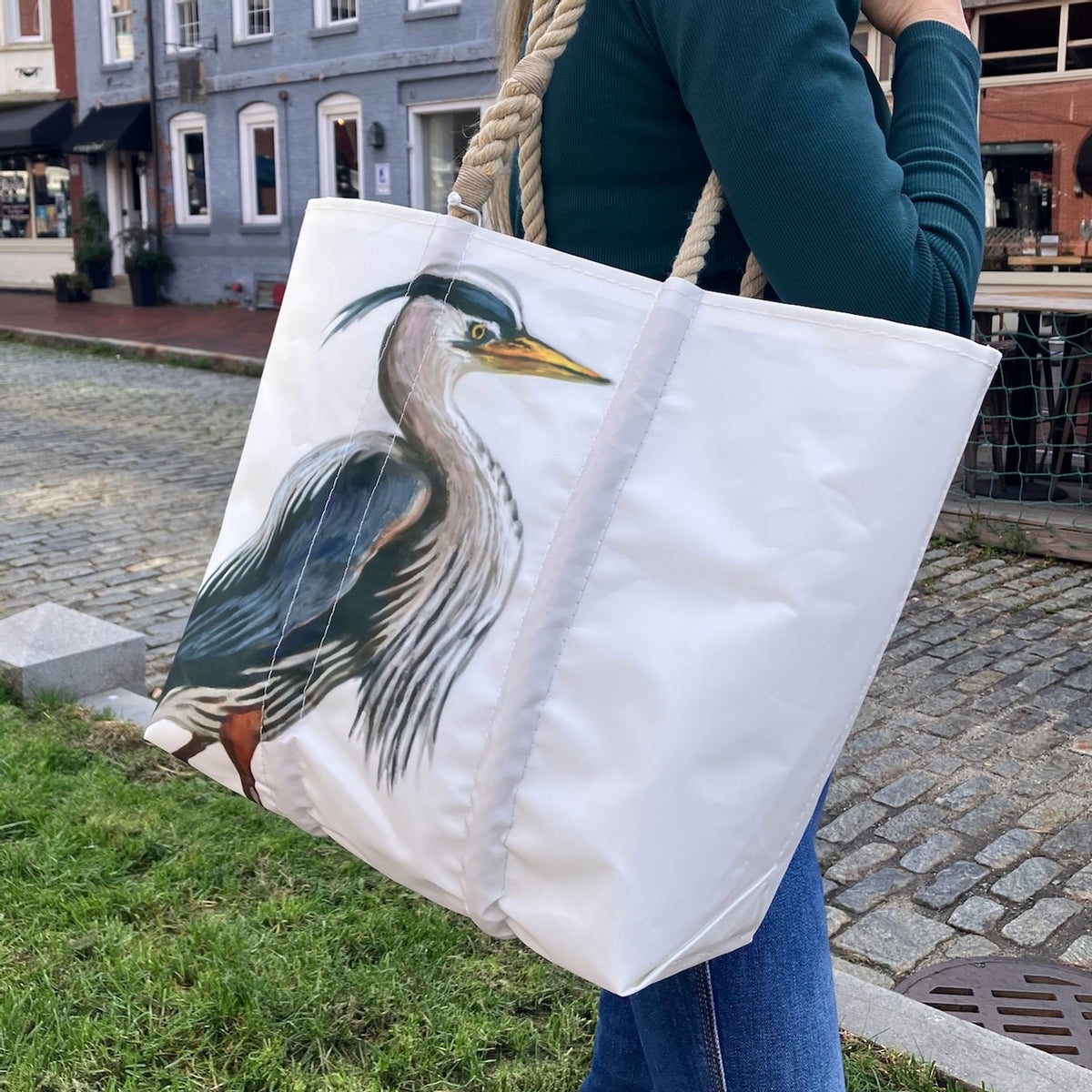 A person stands on grass near a cobblestone street and brick buildings, carrying the Sea Bags Great Blue Heron Medium Tote made from recycled sail cloth.