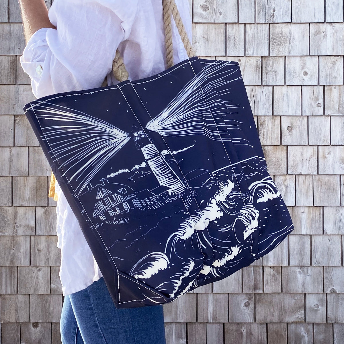 A person in a white shirt carries the Sea Bags Lighthouse Beacon Medium Tote with a seascape, inspired by Portland Head Light, standing before a shingled wall.