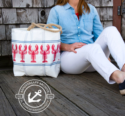 A white Sea Bags Lobster Bake tote with a repeating red lobster print and rope handles, sitting on a wooden deck next to a person.