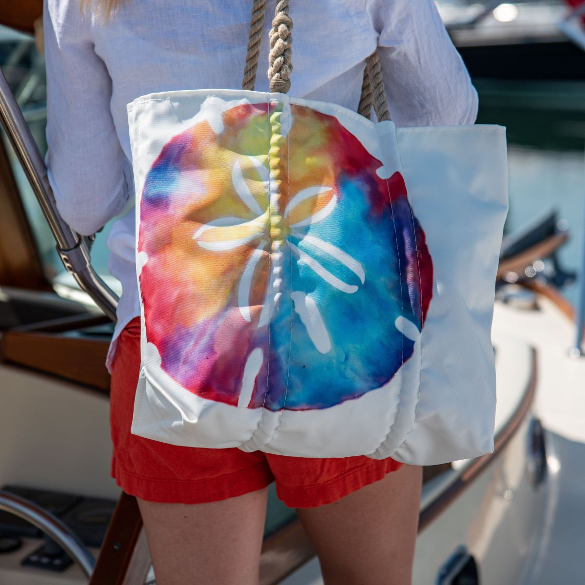 A person in red shorts and a white shirt carries the Sea Bags Tie-Dye Sand Dollar Tote while standing on a boat.