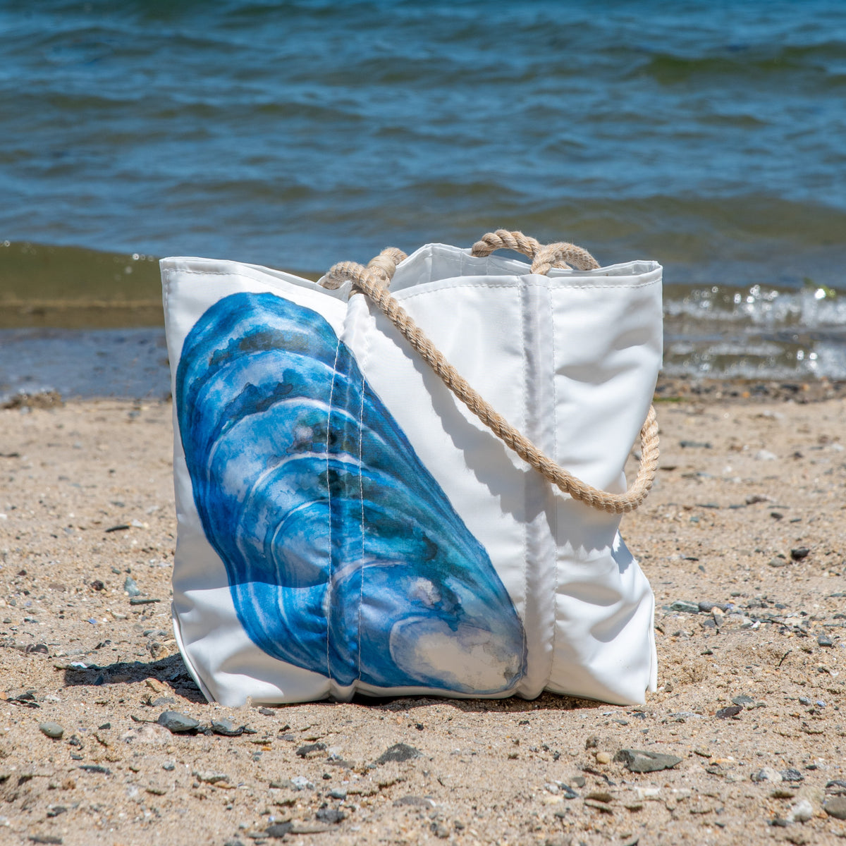 The Mussel Shell Medium Tote by Sea Bags, featuring a blue seashell design and rope handles, rests on sandy beach near shoreline waves.