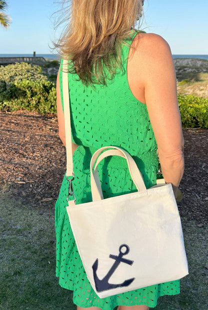 A woman in a green sleeveless dress stands outdoors, carrying the Sea Bags Navy Anchor Crossbody Tote—an eco-friendly bag made from recycled sailcloth and featuring a dark blue anchor design. by Sea Bags