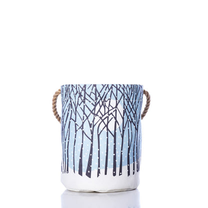 Introducing the Winter Solstice Bucket Bag by Sea Bags—a sustainable basket crafted from recycled sail cloth. This fabric storage basket features sturdy rope handles and showcases a blue and white winter forest design with snow-covered trees set against a crisp white background, making it the perfect stylish solution for seasonal organization. by Sea Bags