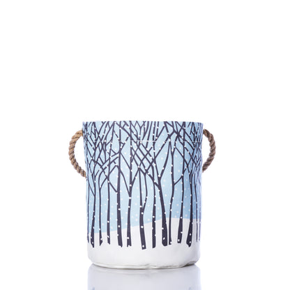 The Sea Bags Winter Solstice Bucket Bag is a cylindrical fabric storage basket made from recycled sail cloth for eco-friendly appeal. Showcasing a winter forest design with leafless trees against a blue backdrop and white snow along the base, it features two tan rope handles. The bag is displayed isolated on a white background. by Sea Bags
