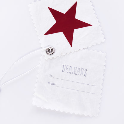 A set of five white sustainable gift tags from Sea Bags, crafted from recycled sail cloth and featuring zigzag edges—each tag includes a red star design or the SEA BAGS MAINE logo with To: and From: blanks, all connected by a metal grommet and string. by Sea Bags