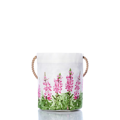 Sea Bags Pink Lupine Bucket features white fabric and sturdy rope handles, accented by a pink and green floral pattern near the base. Shown on a reflective surface with a white background, this stylish bucket adds a fresh touch of spring décor to any room. by Sea Bags