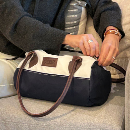 A person sits on a couch, unzipping the Sea Bags Chebeague Baguette Bag, which is made from blue and white recycled sailcloth and features brown handles. by Sea Bags