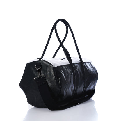 The Sea Bags Black Dragon Duffel, crafted from water-resistant recycled sail cloth, features rope handles and an adjustable shoulder strap. Photographed on a white background with a light reflection, this black and white travel bag combines durability with distinctive maritime style. by Sea Bags
