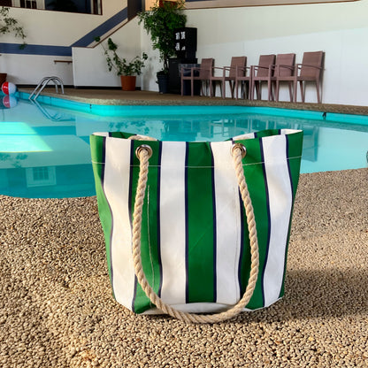 The Sea Bags Preppy Stripe Essential Handbag, featuring classic collegiate green and white stripes and sturdy rope handles, crafted from recycled sailcloth, rests on gravel near an indoor swimming pool with chairs in the background. by Sea Bags