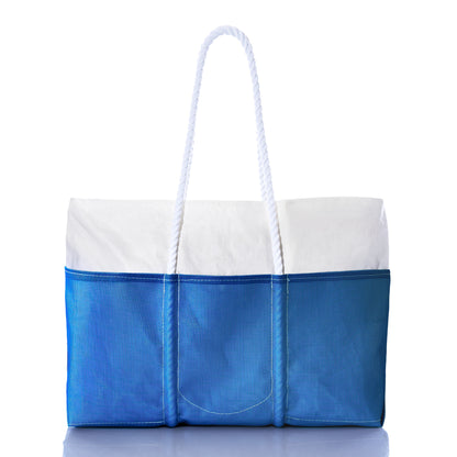 The Sea Bags Harbor Blue Beachcomber Tote—a rectangular beach bag featuring a white top, blue bottom, exterior pockets, and two white rope handles—is displayed against a white background. by Sea Bags