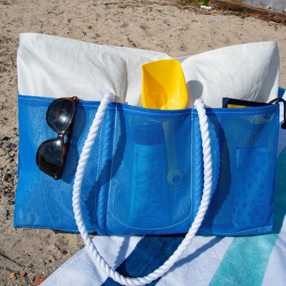 The Sea Bags Harbor Blue Beachcomber Tote, featuring white rope handles and sand-proof mesh construction, holds a white towel, a yellow plastic shovel, bottles, and sunglasses. It’s pictured resting on a beach towel at the shoreline. by Sea Bags
