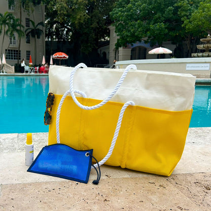A Goldenrod Beachcomber Tote from Sea Bags, featuring sturdy rope handles and a yellow-and-white design, sits poolside alongside sunglasses, sunscreen, and a blue mesh pouch, with umbrellas, lounge chairs, and trees visible in the background. by Sea Bags