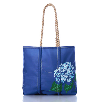 The Harbor Blue Hydrangea Medium Tote by Sea Bags features rope handles and a painted blue hydrangea flower design on the front, displayed against a white background—this charming tote evokes a refreshingly coastal feeling, perfect for sunny days by the shore. by Sea Bags
