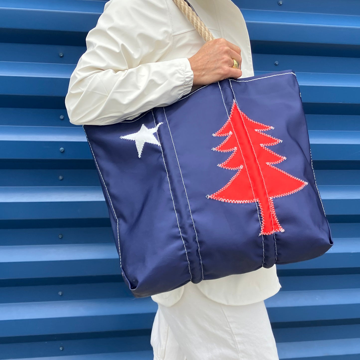 Person holding a blue tote bag with a maine flag red tree and white star design against a blue corrugatedbackground. She wears a waterproof white raincoat. 