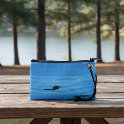 The Sea Bags Life is Good Dog Days Wristlet, featuring a blue design with a black dog graphic, rests on a wooden picnic table by a lakeside surrounded by trees—making it the perfect eco-friendly handbag or wristlet for any adventure. by Sea Bags