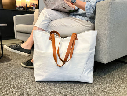 A person sits on a couch reading a newspaper, with the Sea Bags Large Market Tote—featuring brown leather handles—placed on the floor nearby. by Sea Bags