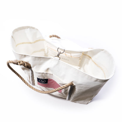 Introducing the Sea Bags U.S. Yacht Ensign Wharf Tote—a white canvas tote featuring rope handles, a metal clasp, and an embroidered American flag patch on the front, combining nautical style with sustainable craftsmanship. by Sea Bags