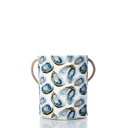 The Watercolor Oyster Bucket by Sea Bags is a white cylindrical container made from recycled sail cloth, adorned with an eye-catching blue watercolor oyster print and finished with rope handles on both sides—capturing the coastal charm of a bucket bag against a crisp white background. by Sea Bags