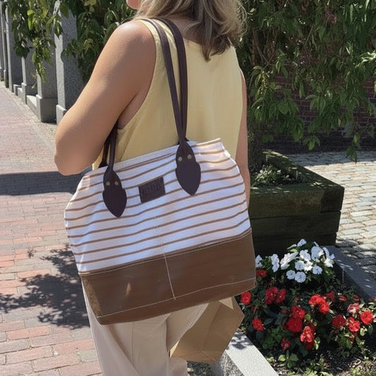 A woman carrying a striped tote bag with brown leather-like handles and a solid brown bottom section, photographed outdoors on a sunny day.