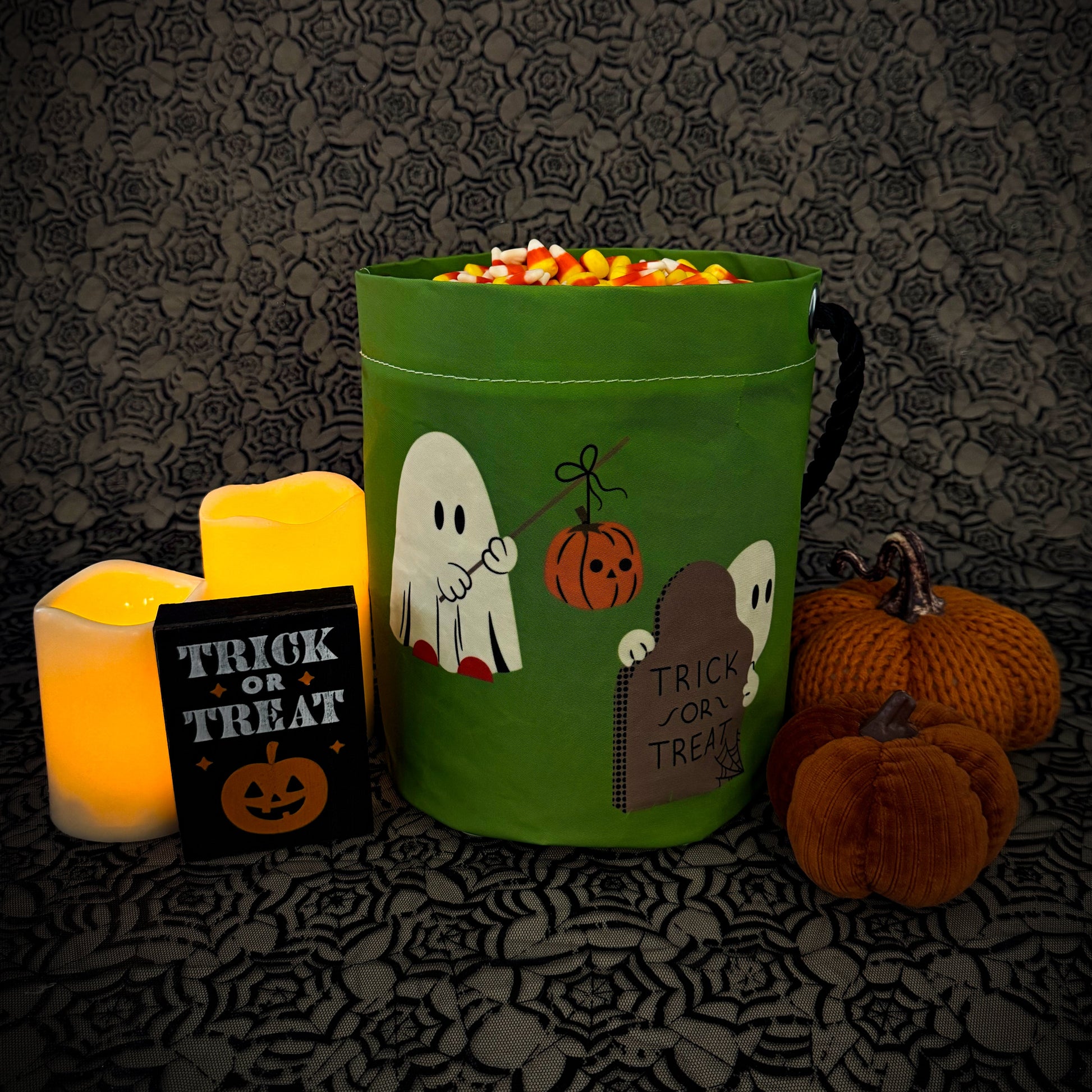 A green fabric Halloween candy bucket with ghost and pumpkin illustrations, filled with candy, styled on a dark patterned surface with candles, a small 'Trick or Treat' sign, and pumpkins.
