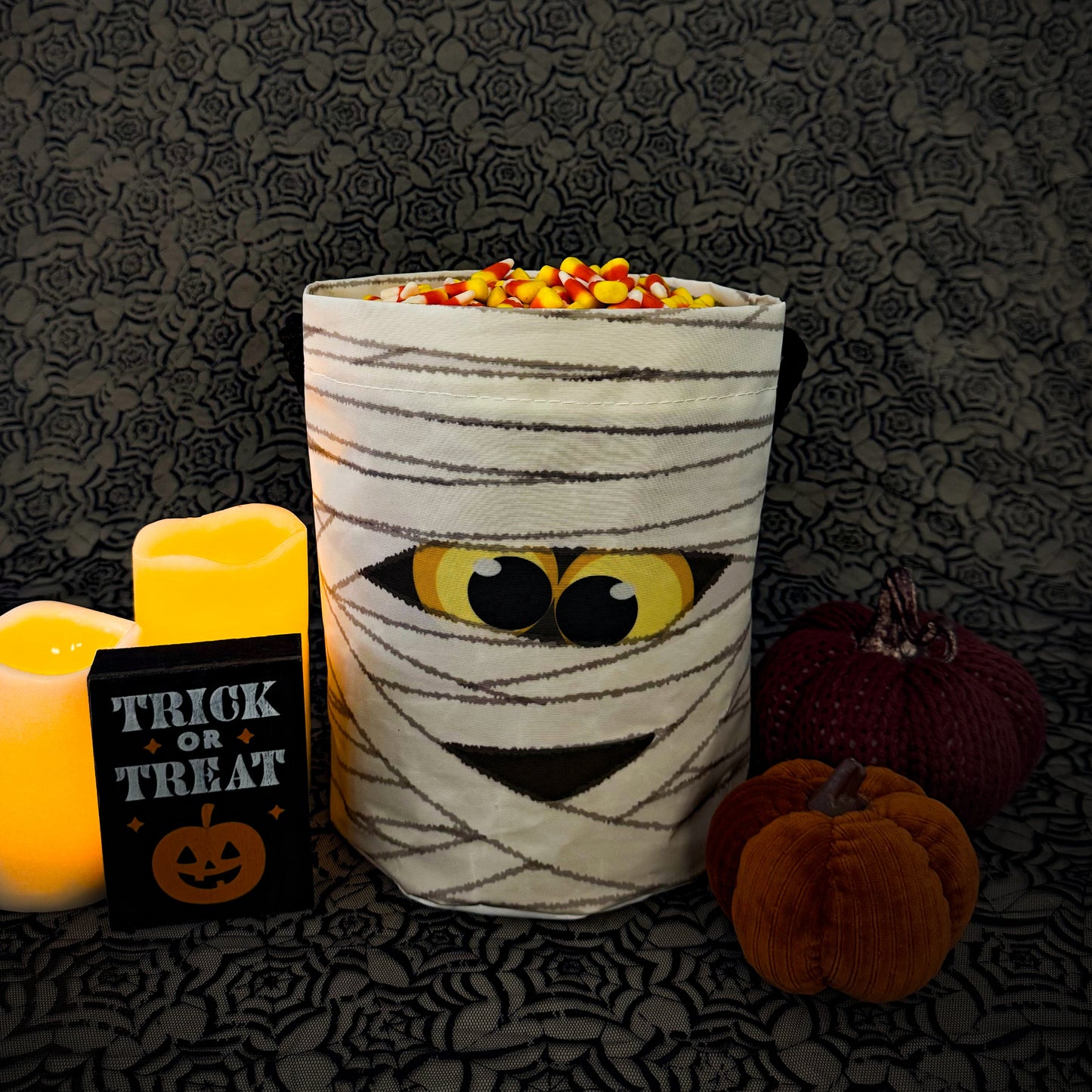 A Halloween-themed mummy decoration made from a white candlestick wrapped in black tape, with candy corn on top and a cartoon eye patch. It is styled with two lit candles, a 'Trick or Treat' sign, and small pumpkins on a dark patterned background.