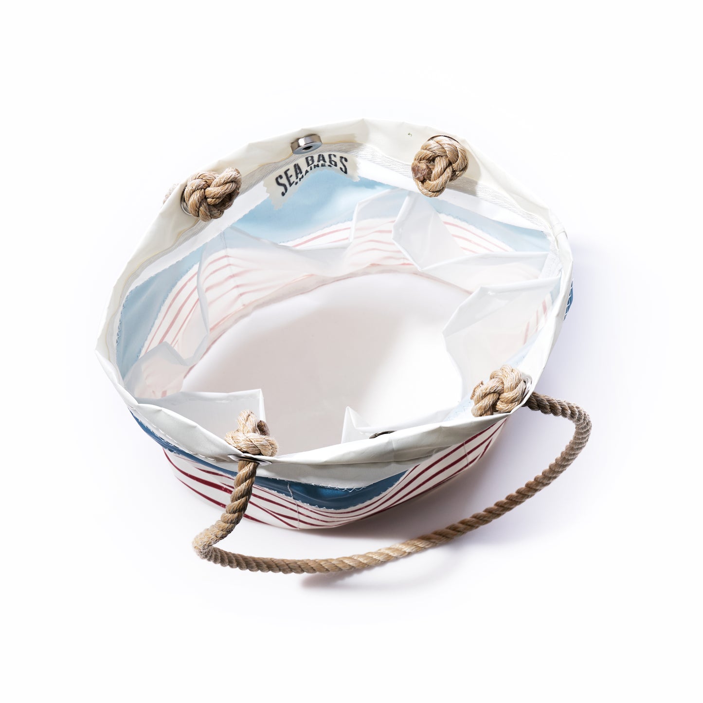 A round essential handbag by Sea Bags, featuring denim blue-on-red mariner stripes, crafted from recycled sail cloth with sturdy rope handles and “SEABASS” printed inside the rim, displayed on a white background.