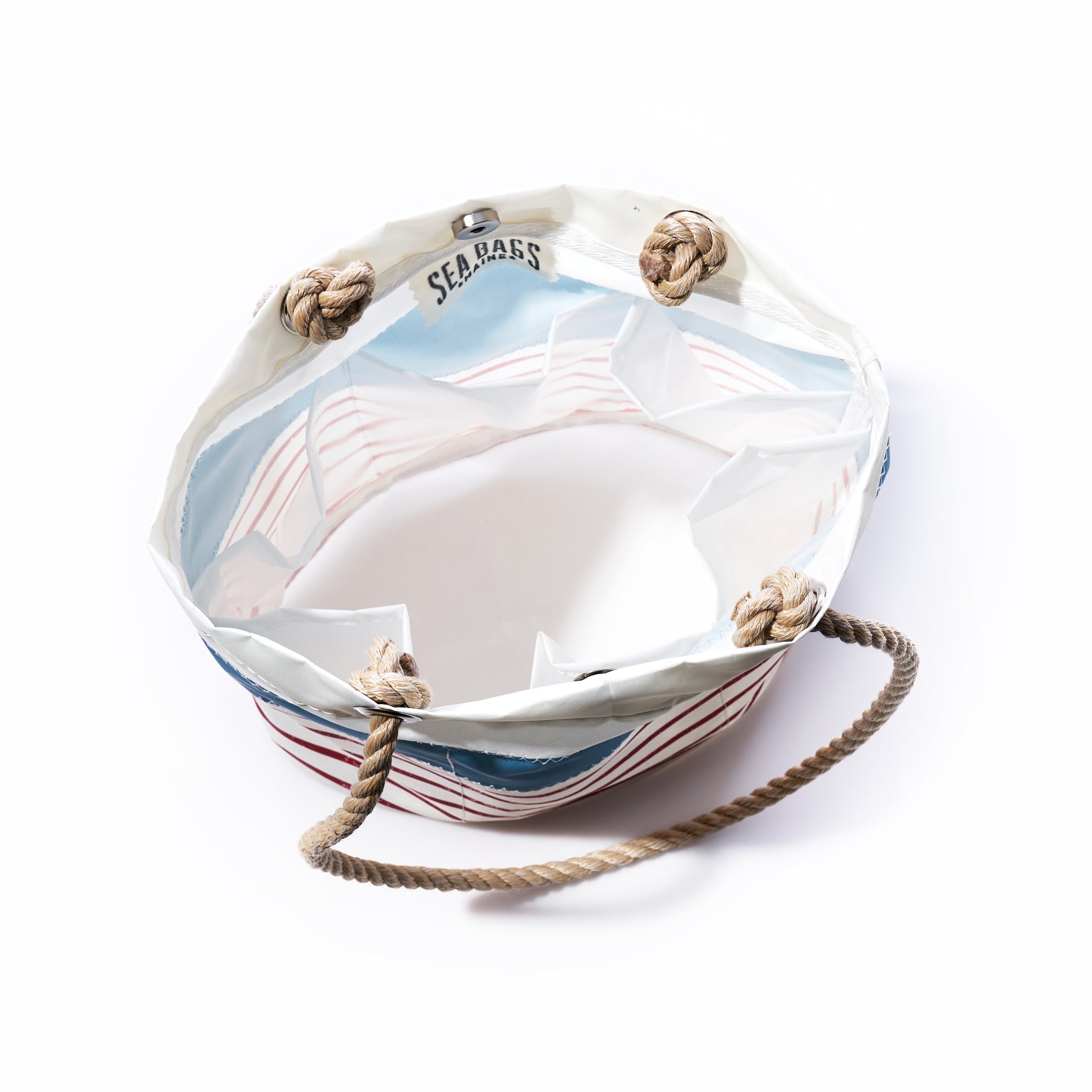 A round essential handbag by Sea Bags, featuring denim blue-on-red mariner stripes, crafted from recycled sail cloth with sturdy rope handles and “SEABASS” printed inside the rim, displayed on a white background.