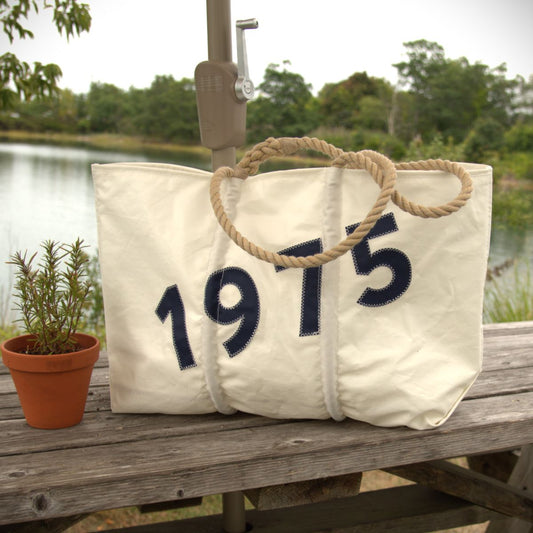 A white Sea Bags Custom Year Large Tote with rope handles and “1975” in blue rests on a wooden bench beside a small potted plant, with a lake and trees visible in the background.