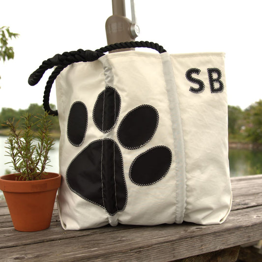 The Sea Bags Custom Monogram Paw Print Medium Tote features black rope handles, a black paw print design, and “SB” monogramming. Shown outdoors on a wooden surface beside a small potted plant, this stylish tote is perfect for everyday use and makes a thoughtful gift for dog lovers.