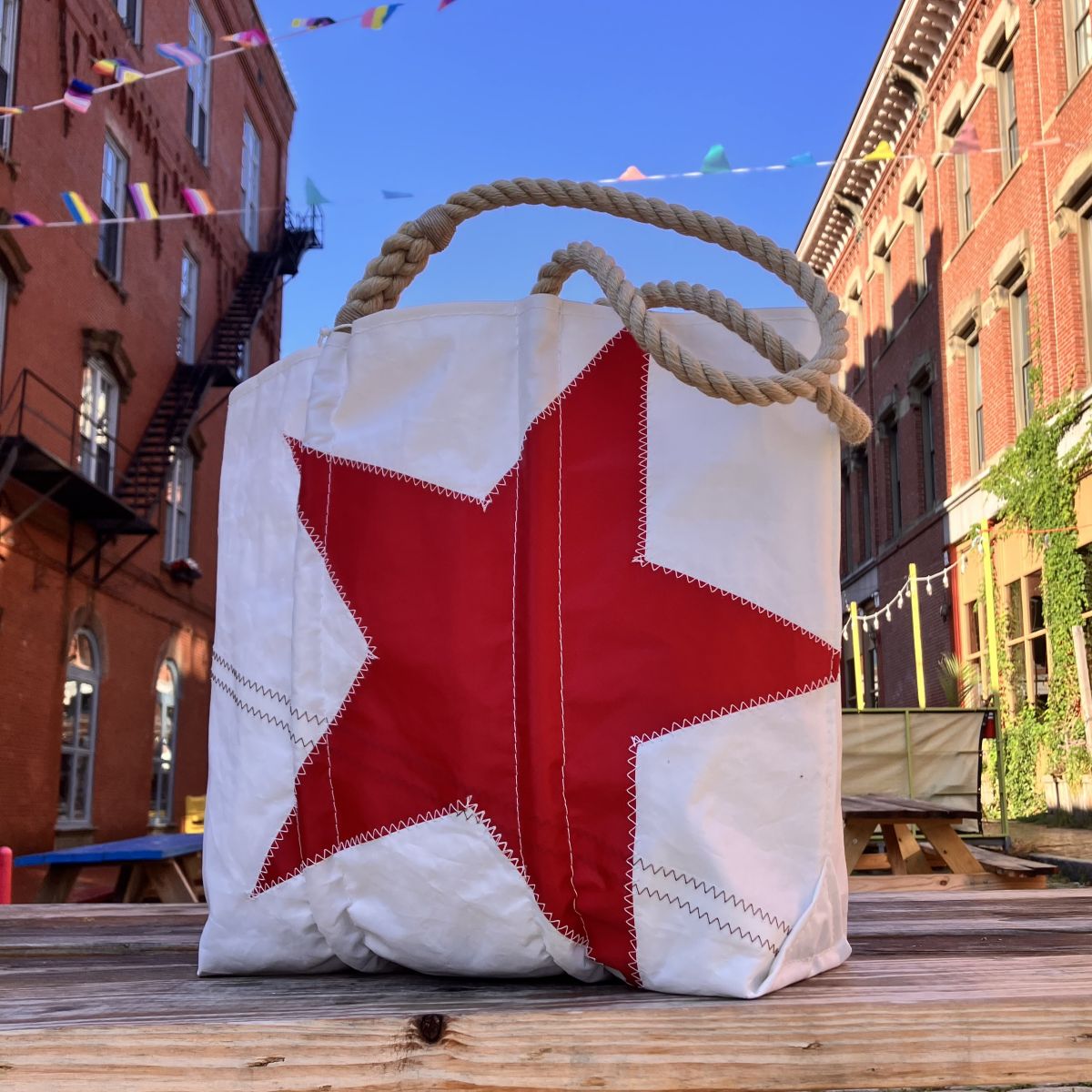 The Sea Bags Red Star Medium Tote, a white handcrafted bag from the Anniversary Archive Collection with striking red star detailing and sturdy rope handles, is displayed on a wooden table outdoors amid brick buildings and vibrant string flags.