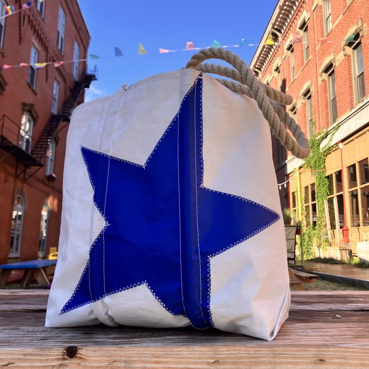 A Royal Blue Star Medium Tote by Sea Bags—featuring a crisp white body with a bold blue star and sturdy rope handles—rests on a wooden table outdoors, with brick buildings and vibrant flags in the background. Handcrafted for both style and utility, this bag is the perfect companion for any outing.