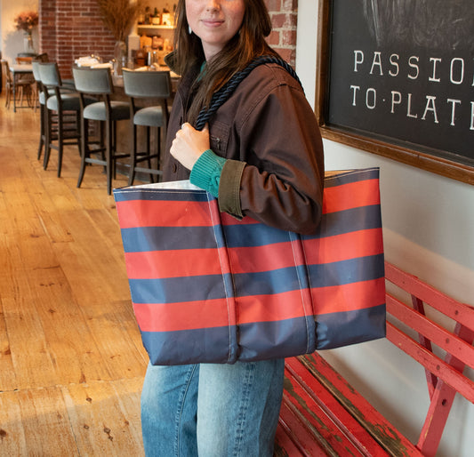 A woman in a brown jacket and blue jeans carries a large striped tote bag with horizontal navy blue and coral red stripes, standing in a rustic cafe setting.