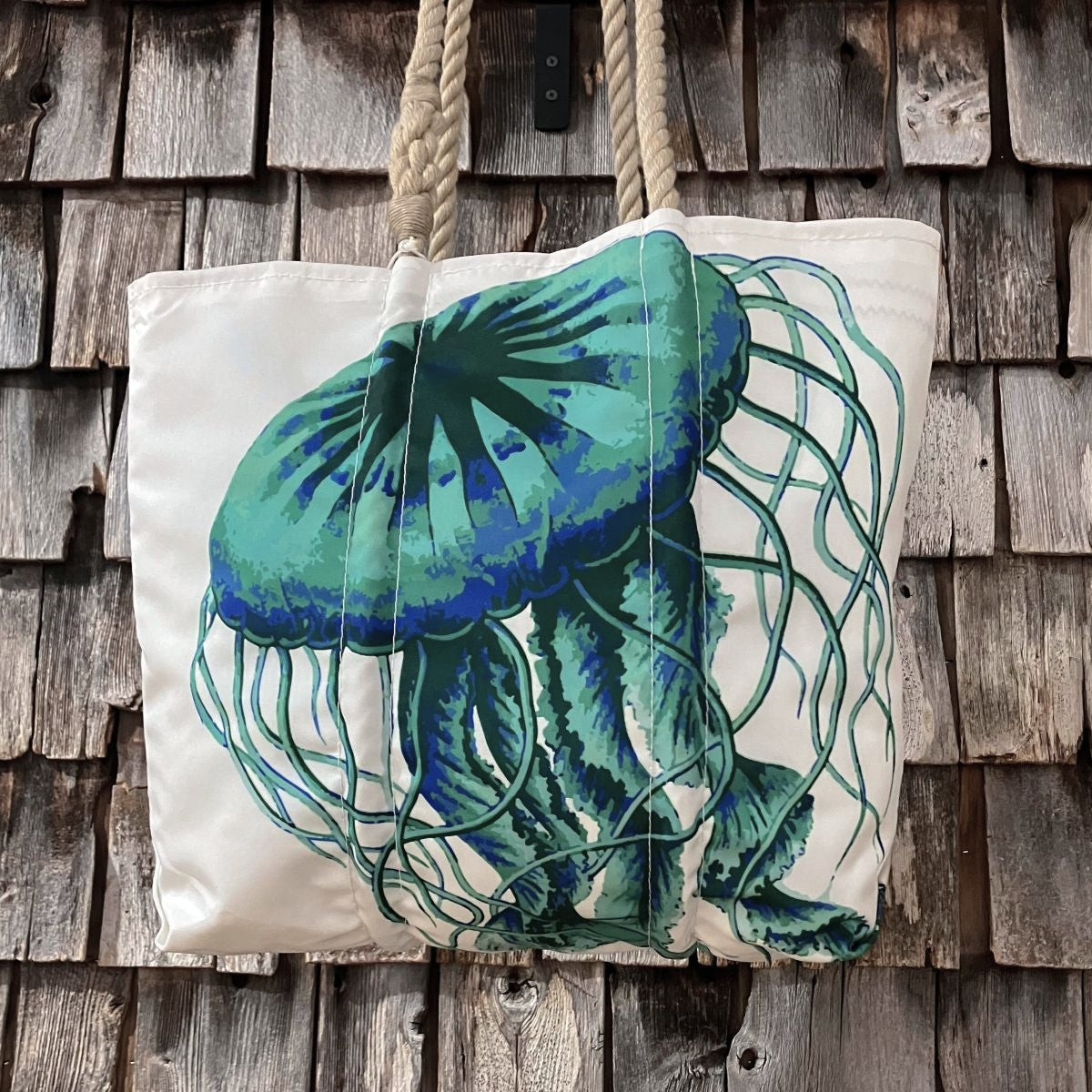 Jellyfish Medium Tote white canvas bag with a blue and green jellyfish design and rope handles, hanging against a weathered wood shingle wall.