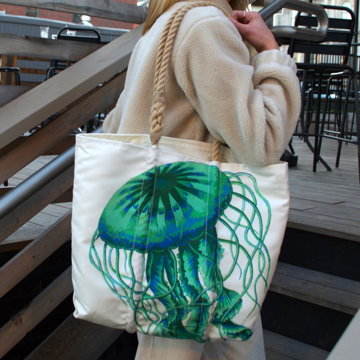 A person in a light-colored fleece jacket carries a Jellyfish Medium Tote by Sea Bags, featuring a large green and blue jellyfish graphic and rope handles.