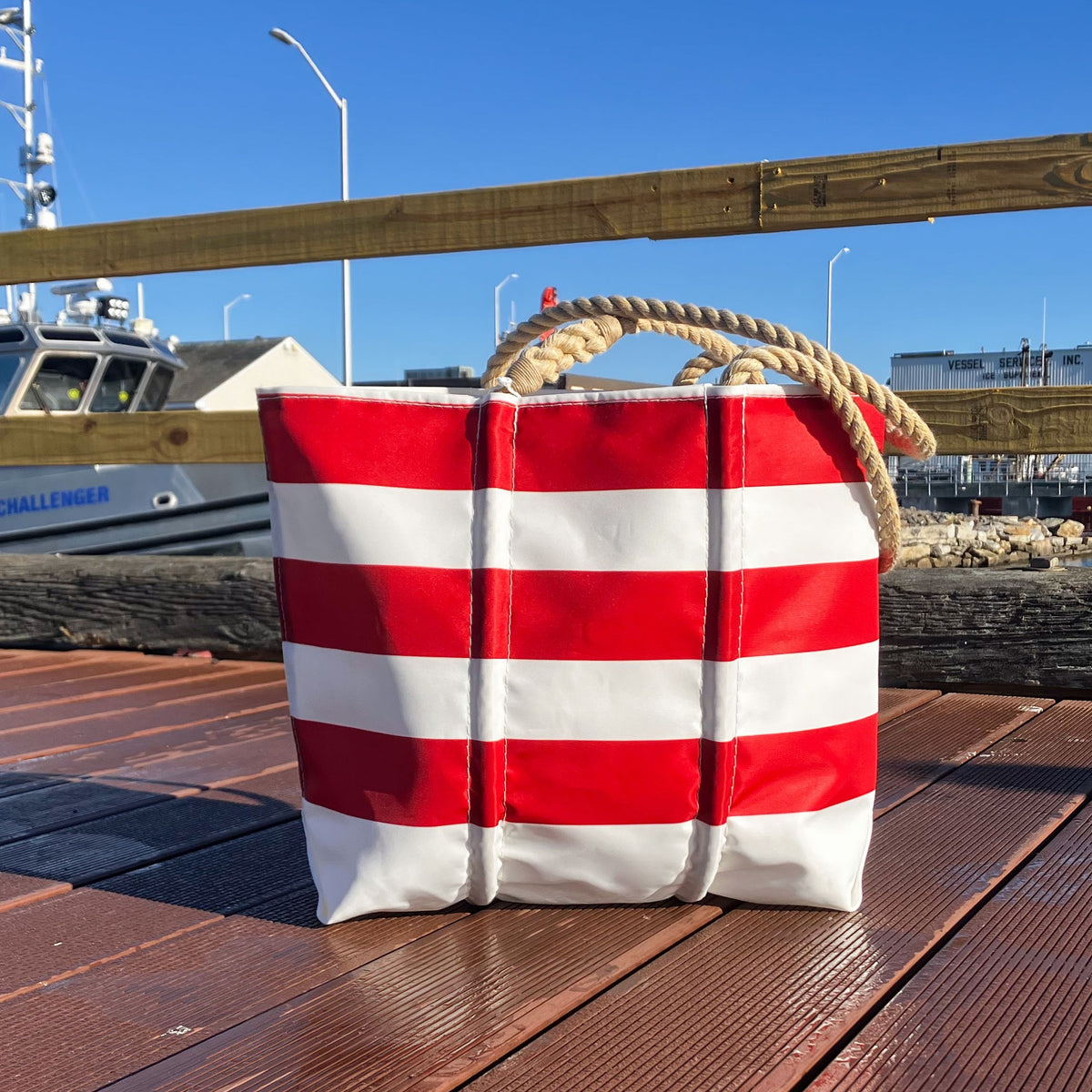 A Red Rugby Stripe Medium Tote by Sea Bags with red and white horizontal lines and thick rope handles, sitting on a wooden dock.