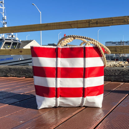 A Red Rugby Stripe Medium Tote by Sea Bags with red and white horizontal lines and thick rope handles, sitting on a wooden dock.