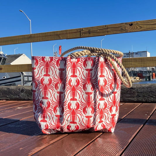 Red Lobster Medallion Medium Tote by Sea Bags with a repeating red lobster pattern and thick rope handles, sitting on a wooden pier.