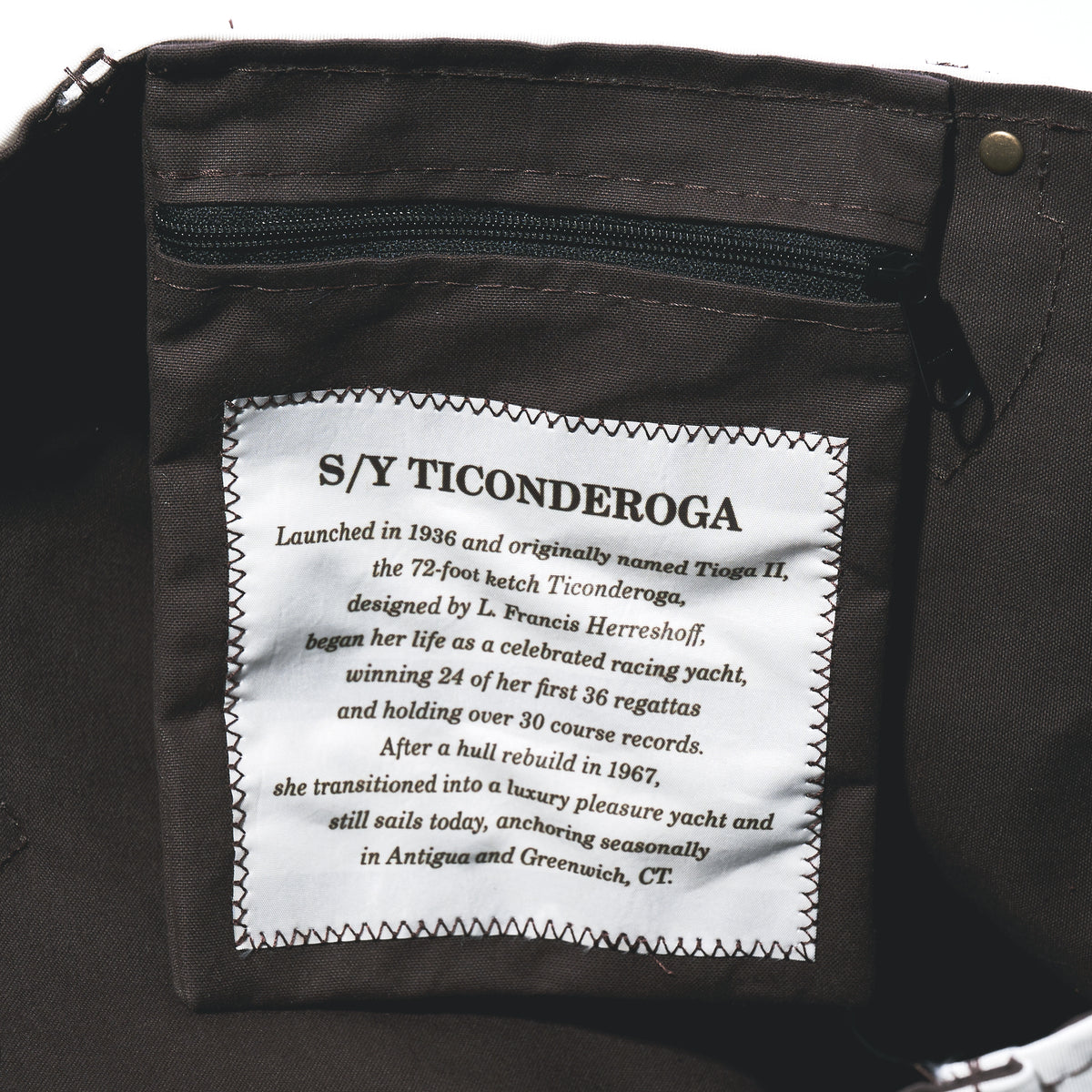 Interior pocket of a dark brown vintage sail cloth Heritage Ticonderoga Medium Tote, featuring a white rectangular patch that recounts the history of the yacht S/Y Ticonderoga.