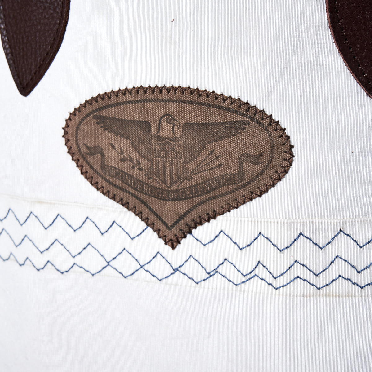 Heritage Ticonderoga Medium Tote in white vintage sail cloth, featuring a brown shield-shaped patch with an eagle emblem and two rows of blue zigzag stitching.
