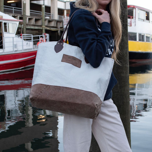 A person carries a Heritage Ticonderoga Medium Tote, a limited edition vintage sail cloth bag with brown leather straps, while standing on a dock next to boats.
