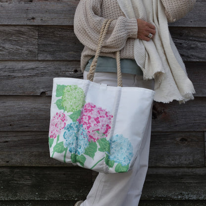 A person wearing a beige sweater holds a 2026 Hope In Bloom Medium Tote with Clasp, a recycled sail cloth bag decorated with green, pink, and blue hydrangea flowers.