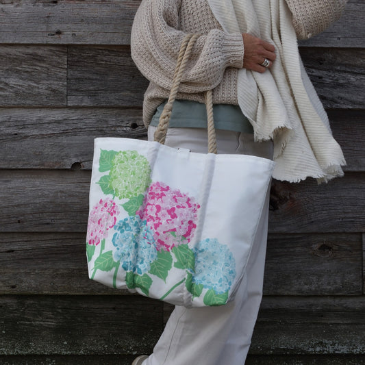 A person wearing a beige sweater holds a 2026 Hope In Bloom Medium Tote with Clasp, a recycled sail cloth bag decorated with green, pink, and blue hydrangea flowers.