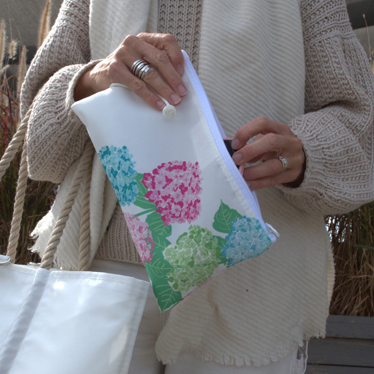 A person holds a white recycled sail cloth 2026 Hope In Bloom Large Wristlet featuring a print of pink, blue, and green hydrangea flowers.
