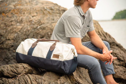 A person sits on rocks by the water, wearing a gray polo and blue pants, with the Sea Bags Chebeague Duffel—crafted from recycled sail and featuring premium leather handles—in white and navy beside them. by Sea Bags