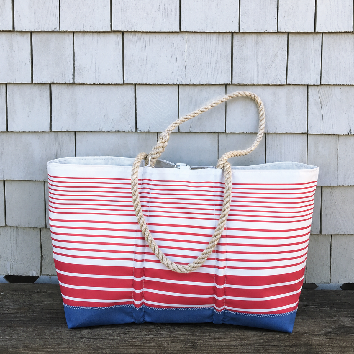 The Red Stripe Ogunquit Beach Tote by Sea Bags—a large, limited edition bag featuring red and white horizontal stripes, blue bottom corners, and thick rope handles—is displayed in front of a shingled wall.