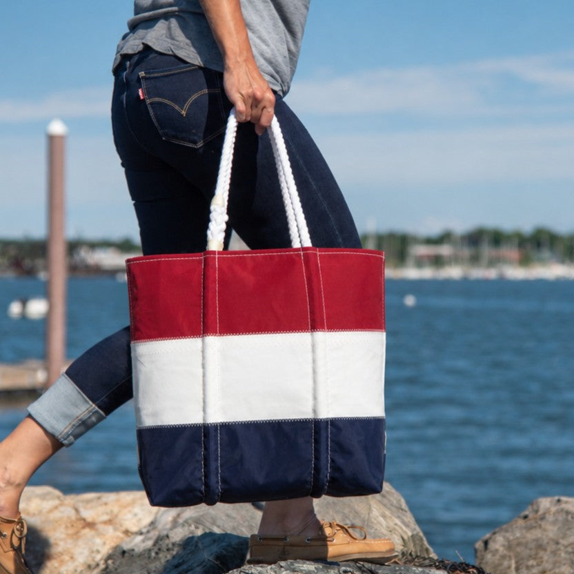 A person carrying the Sea Bags Red White and Navy Stripe Medium Tote with rope handles near a body of water, with a marina in the background.